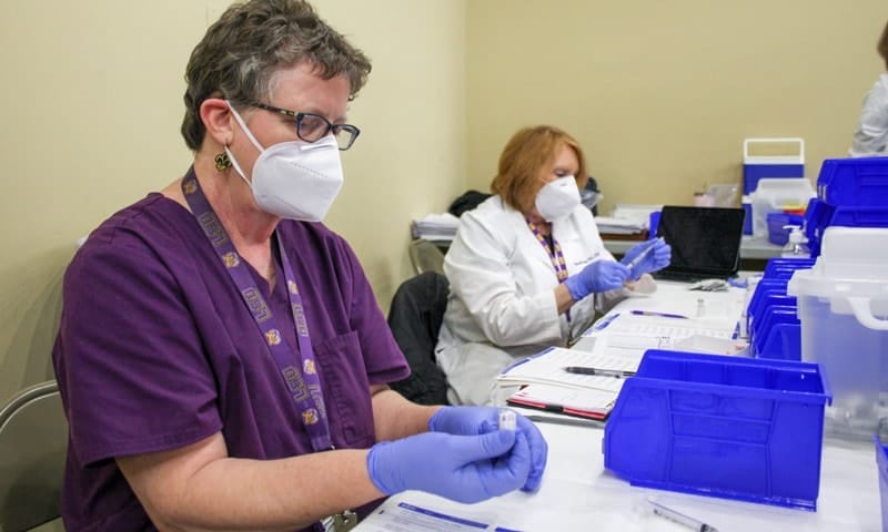 Two vaccination clinic staff members, one in purple scrubs and one in a lab coat, prepare Pfizer vaccine doses.
