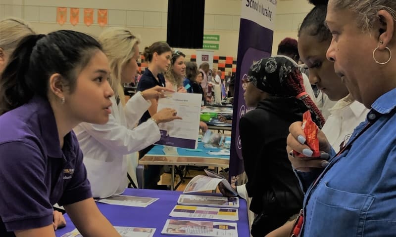 A School of Nursing student in a purple shirt offers recruitment paperwork to a high school student and the woman accompanying her.
