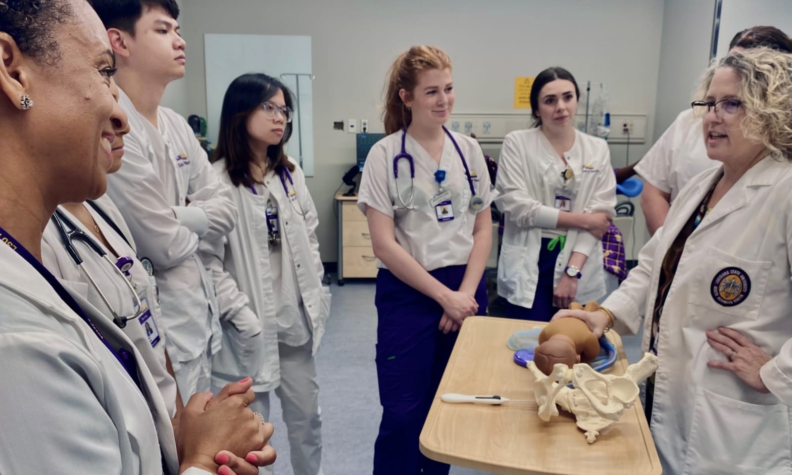 A group of people look on as a nurse midwife in a white coat shows how a baby mannequin fits in the model of a woman’s pelvis.