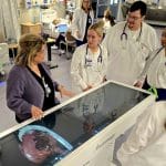 A group of healthcare professionals and students in a clinical setting, gathered around an interactive Anatomage table displaying a 3D anatomical model.