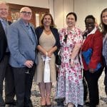 Group of six attendees at an event, smiling and posing together in formal and semi-formal attire, inside a bright room with tables and chairs in the background.