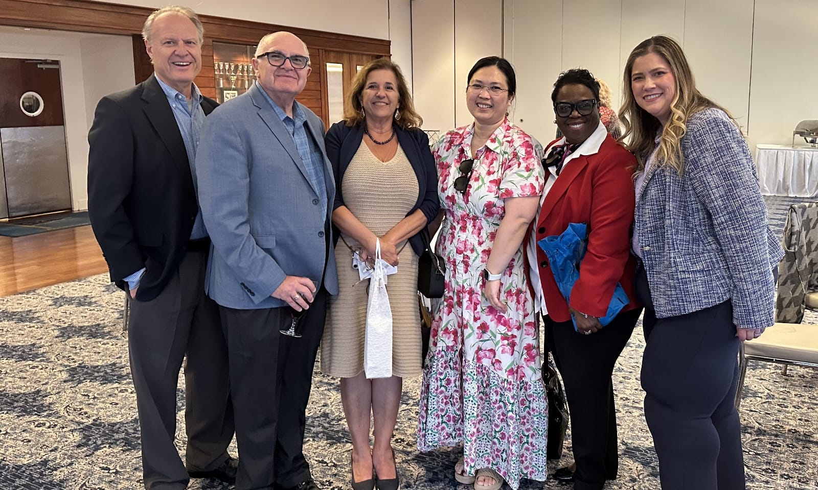 Group of six attendees at an event, smiling and posing together in formal and semi-formal attire, inside a bright room with tables and chairs in the background.