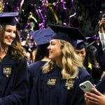 Two LSU Health graduates in caps and gowns smiling at each other during a graduation ceremony, with confetti and other graduates celebrating in the background.